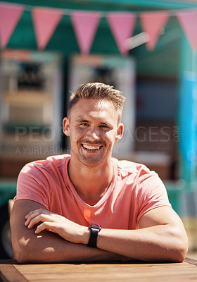 Buy stock photo Portrait of a cheerful man seated at a table by himself outside next to a beach promenade during the day