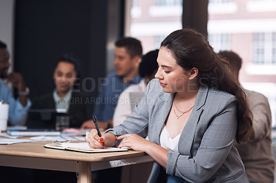 Buy stock photo Shot of a young businesswoman writing in a notebook while sitting in an office with her colleagues in the background