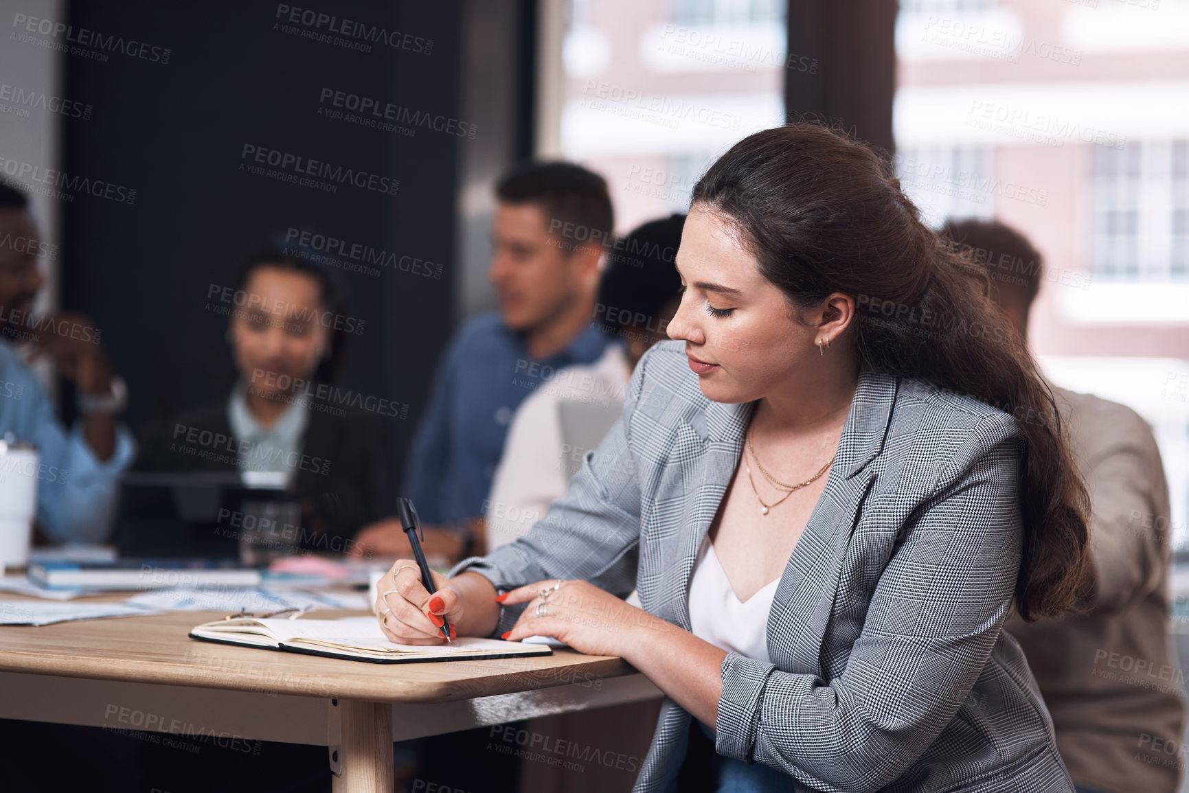 Buy stock photo Shot of a young businesswoman writing in a notebook while sitting in an office with her colleagues in the background