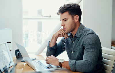 Buy stock photo Cropped shot of a handsome young businessman sitting alone and looking contemplative while using his laptop in the office