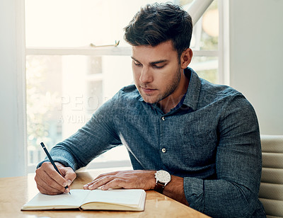 Buy stock photo Cropped shot of a handsome young businessman sitting and writing in a notebook while in the office alone