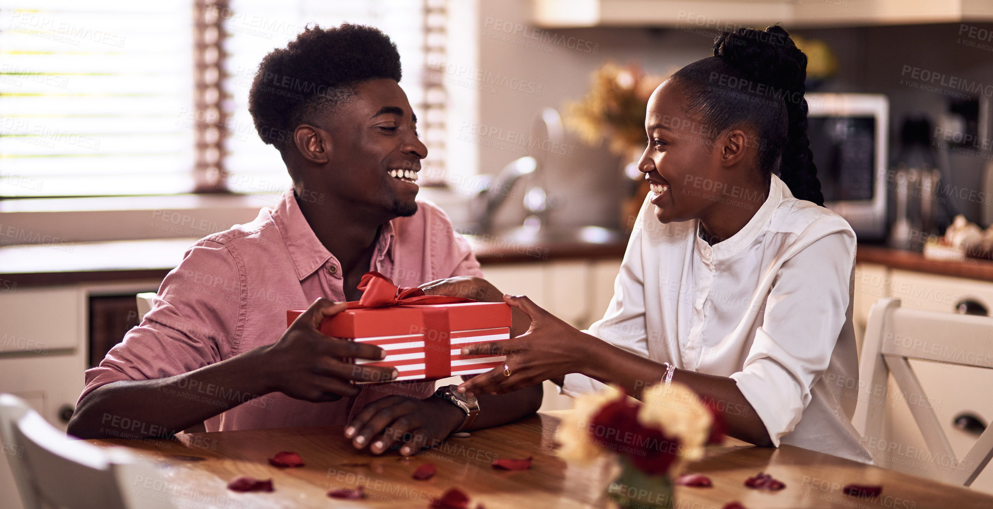 Buy stock photo Black couple, kitchen and present in home with love, smile and bonding together with rose petals. Happy, man and woman holding hands with gift, relax and celebrating Valentines day or anniversary