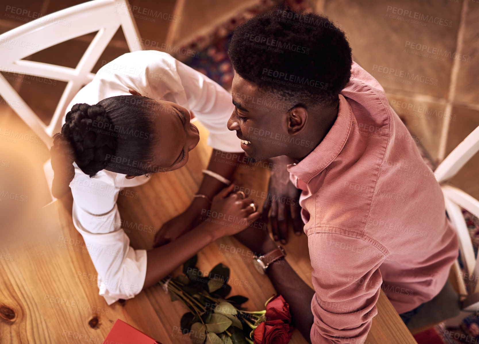 Buy stock photo Holding hands, love and black couple in restaurant for date on valentines day or anniversary. Happy, kiss and African man and woman with connection, bonding or romance at diner with roses from above.