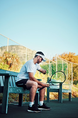 Buy stock photo Shot of a sporty young man checking his watch while sitting on a bench on a tennis court