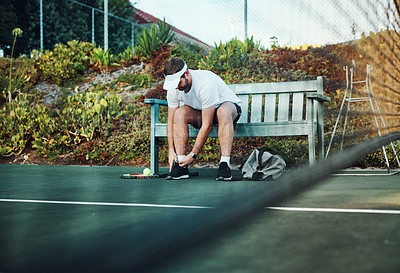 Buy stock photo Shot of a sporty young man tying his laces while sitting on a bench on a tennis court