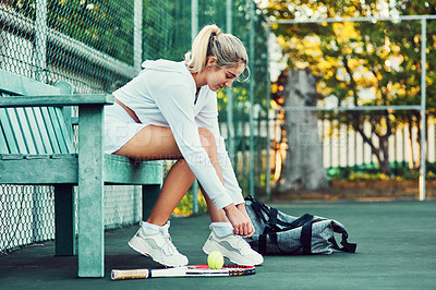 Buy stock photo Shot of a sporty young woman tying her laces while sitting on a bench on a tennis court