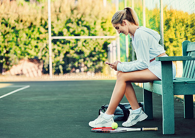 Buy stock photo Shot of a sporty young woman using a cellphone while sitting on a bench on a tennis court