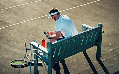 Buy stock photo Shot of a sporty young man using a cellphone while sitting on a bench on a tennis court