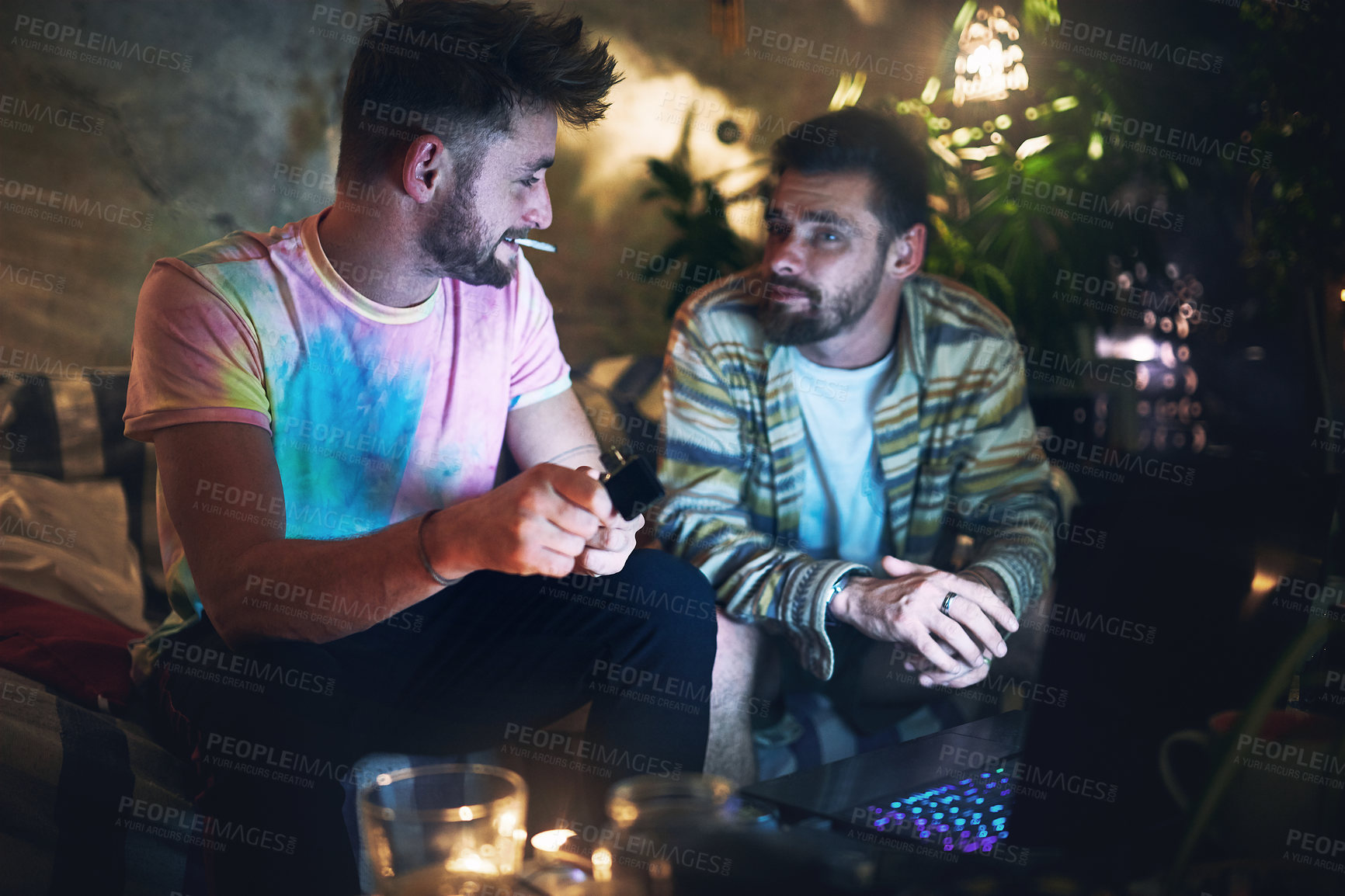Buy stock photo Cropped shot of a handsome young man lighting up a joint of cannabis while sitting with is friend at home