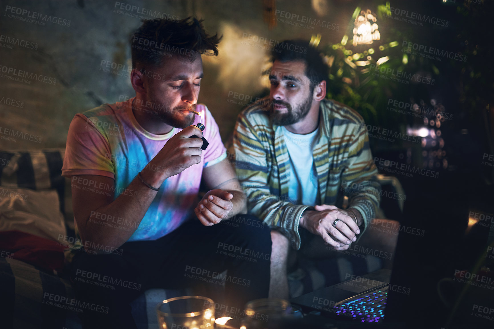 Buy stock photo Cropped shot of a handsome young man lighting up a joint of cannabis while sitting with is friend at home