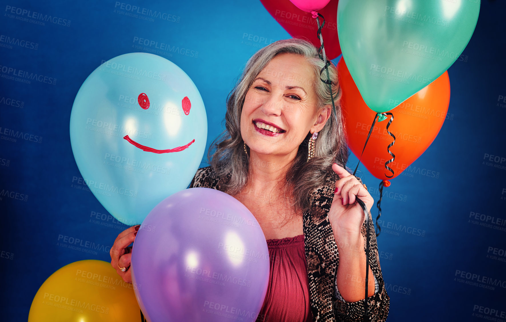Buy stock photo Portrait of a cheerful senior woman posing holding balloons in studio against a blue background