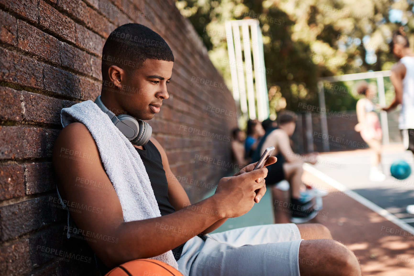 Buy stock photo Shot of a sporty young man using a cellphone while taking a break after a game of basketball