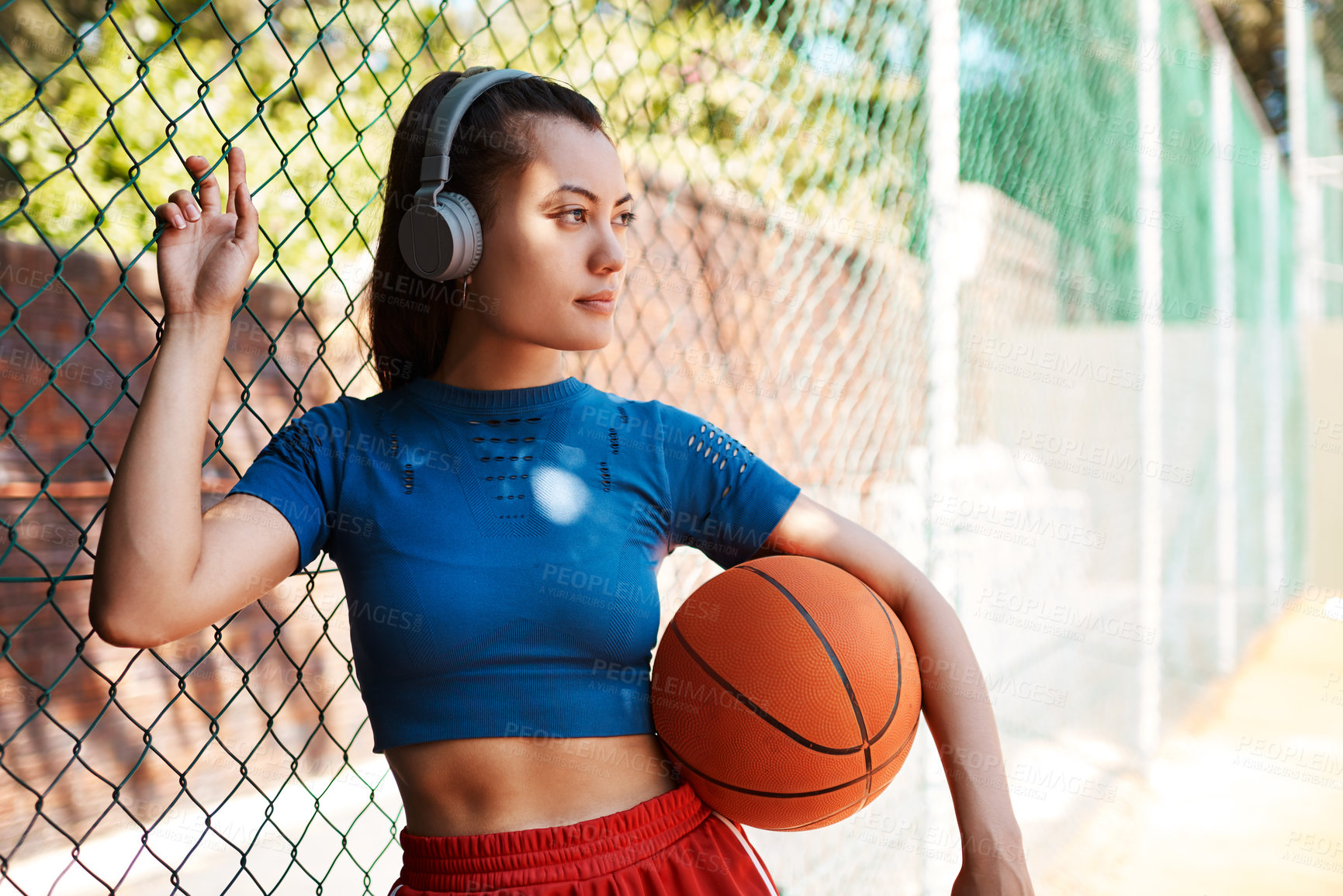 Buy stock photo Shot of a sporty young woman holding a basketball while standing against a fence outdoors