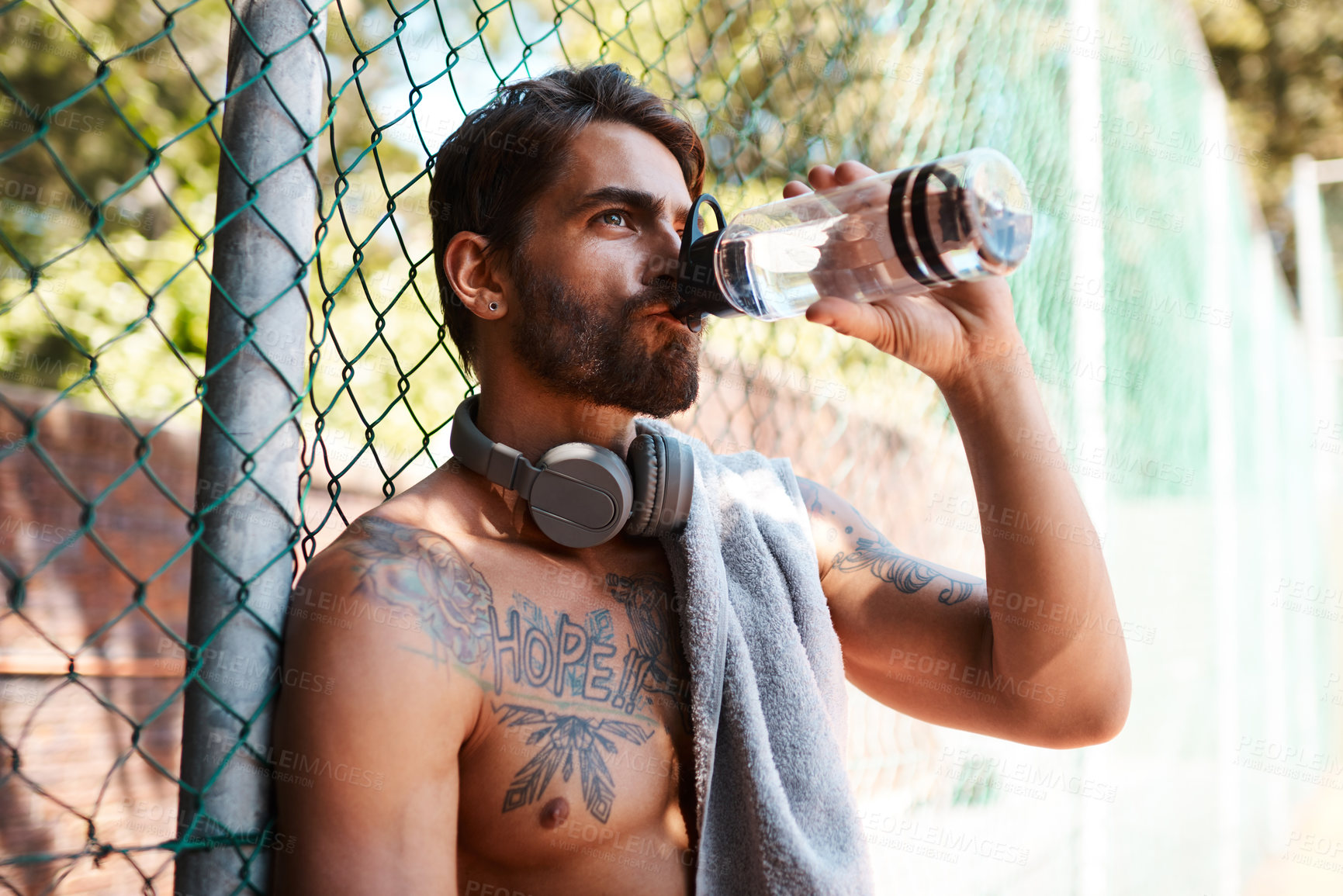 Buy stock photo Shot of a sporty young man drinking water while standing against a fence outdoors