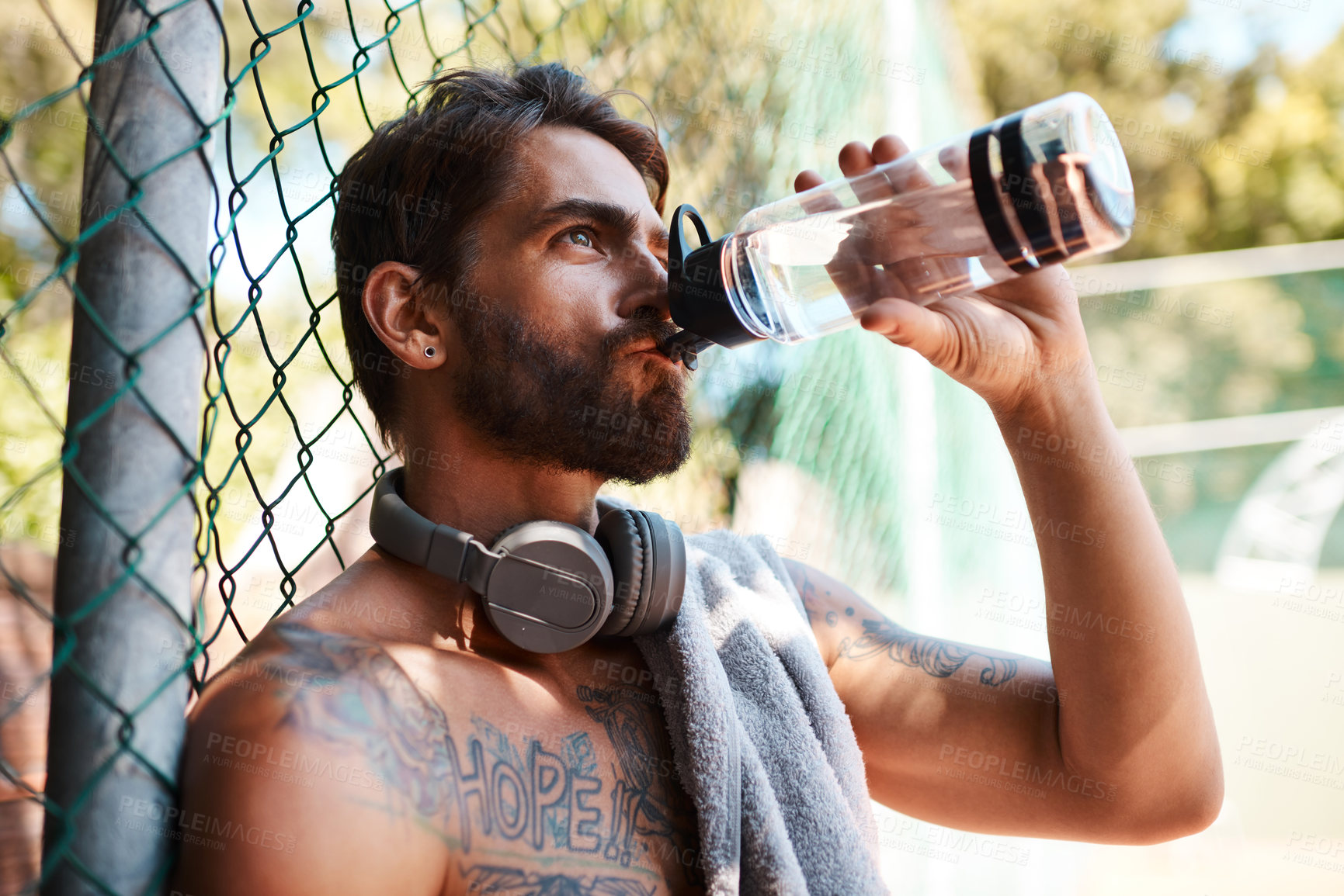 Buy stock photo Shot of a sporty young man drinking water while standing against a fence outdoors