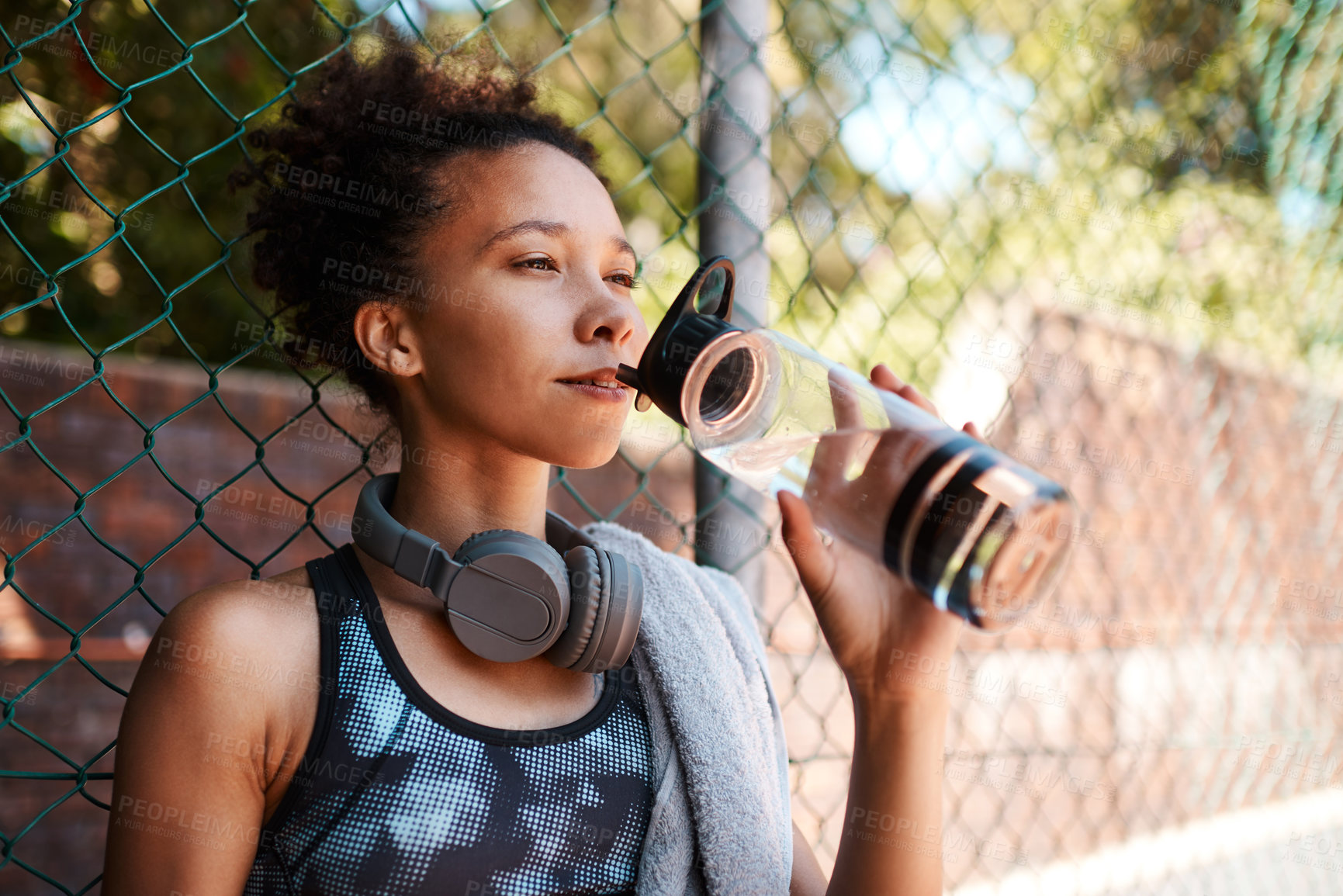Buy stock photo Shot of a sporty young woman drinking water while standing against a fence outdoors