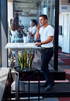 Buy stock photo Shot of a young businessman using a cellphone while walking on a treadmill in an office
