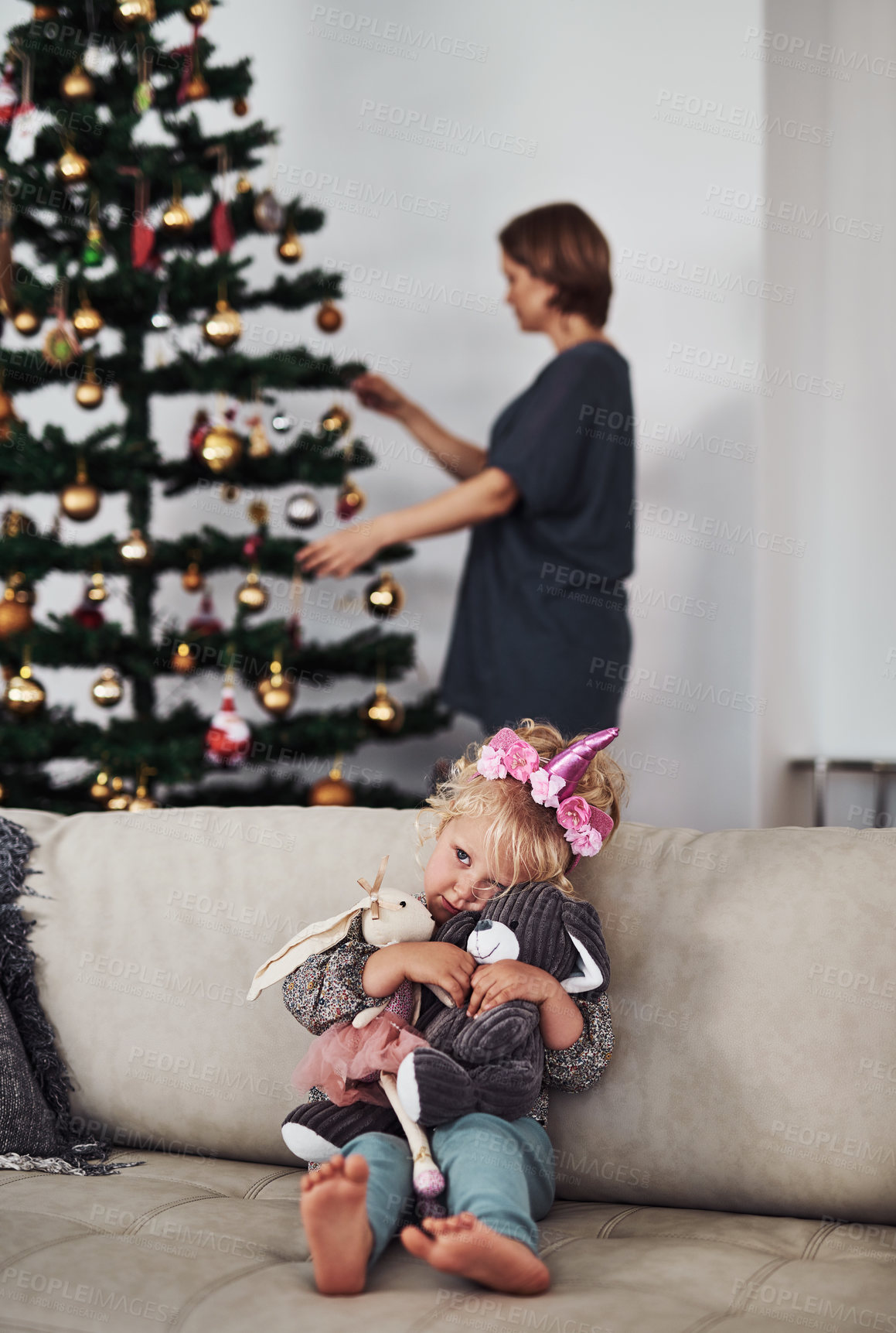 Buy stock photo Cropped shot of an adorable little girl sitting on the sofa while her mother fixes the Christmas tree behind her