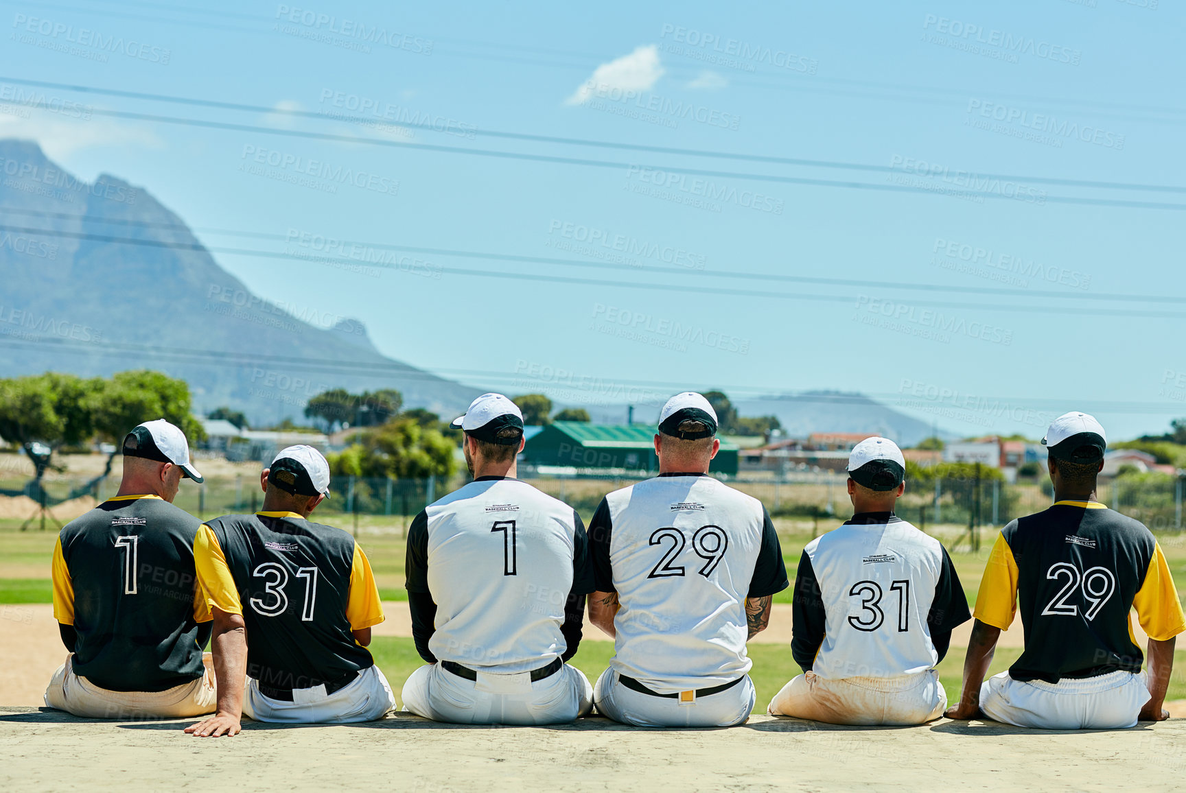 Buy stock photo Rearview shot of a team of unrecognizable baseball players sitting together near a baseball field during the day