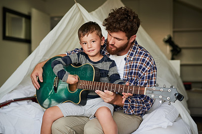 Buy stock photo Cropped portrait of an adorable little boy sitting on his dad's lap while learning to play the guitar at home