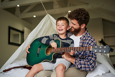 Buy stock photo Cropped portrait of an adorable little boy sitting on his dad's lap while learning to play the guitar at home