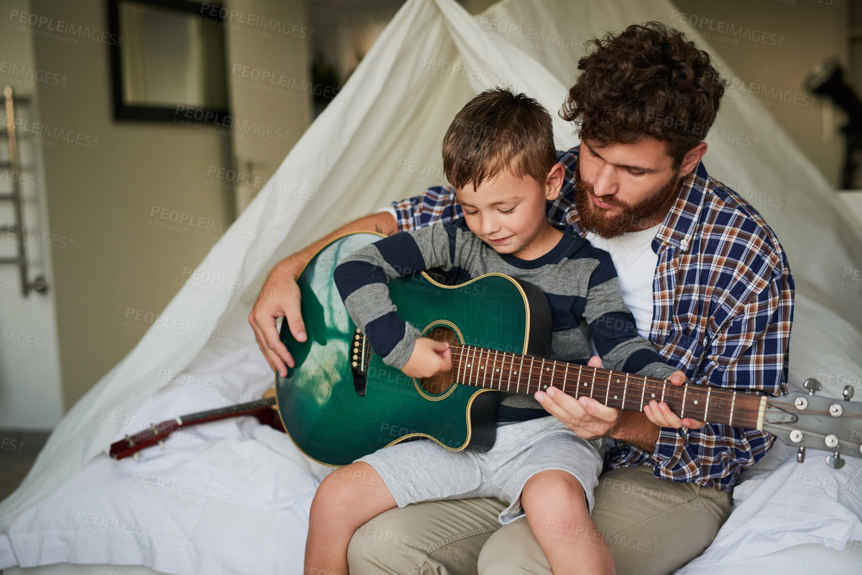 Buy stock photo Cropped shot of an adorable little boy sitting on his dad's lap while learning to play the guitar at home
