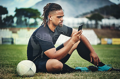 Buy stock photo Full length shot of a handsome young rugby player using a smartphone while sitting in the playing field during the day