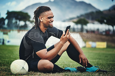 Buy stock photo Full length shot of a handsome young rugby player using a smartphone while sitting in the playing field during the day