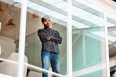 Buy stock photo Shot of a young businessman looking thoughtful while standing at a glass wall in an office
