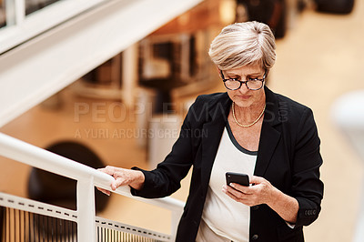 Buy stock photo Shot of a mature businesswoman using a cellphone while walking up a staircase in an office