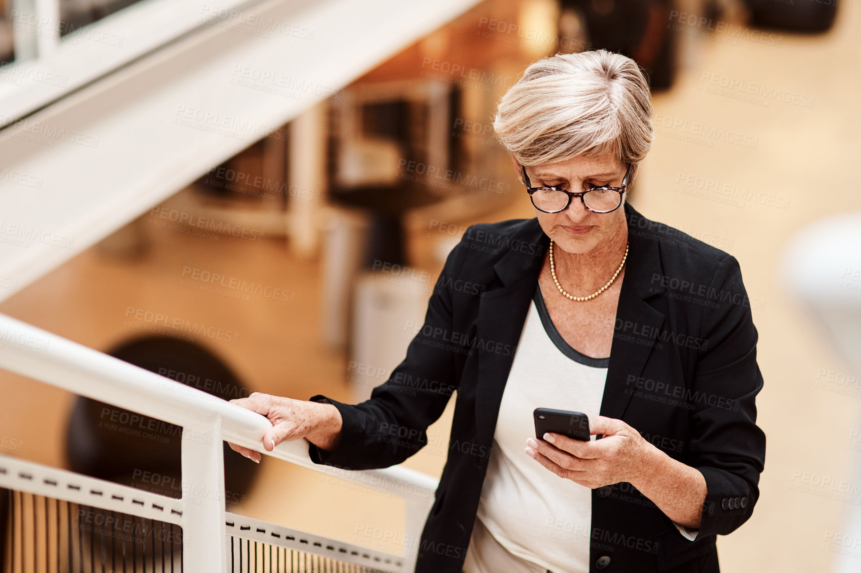 Buy stock photo Shot of a mature businesswoman using a cellphone while walking up a staircase in an office