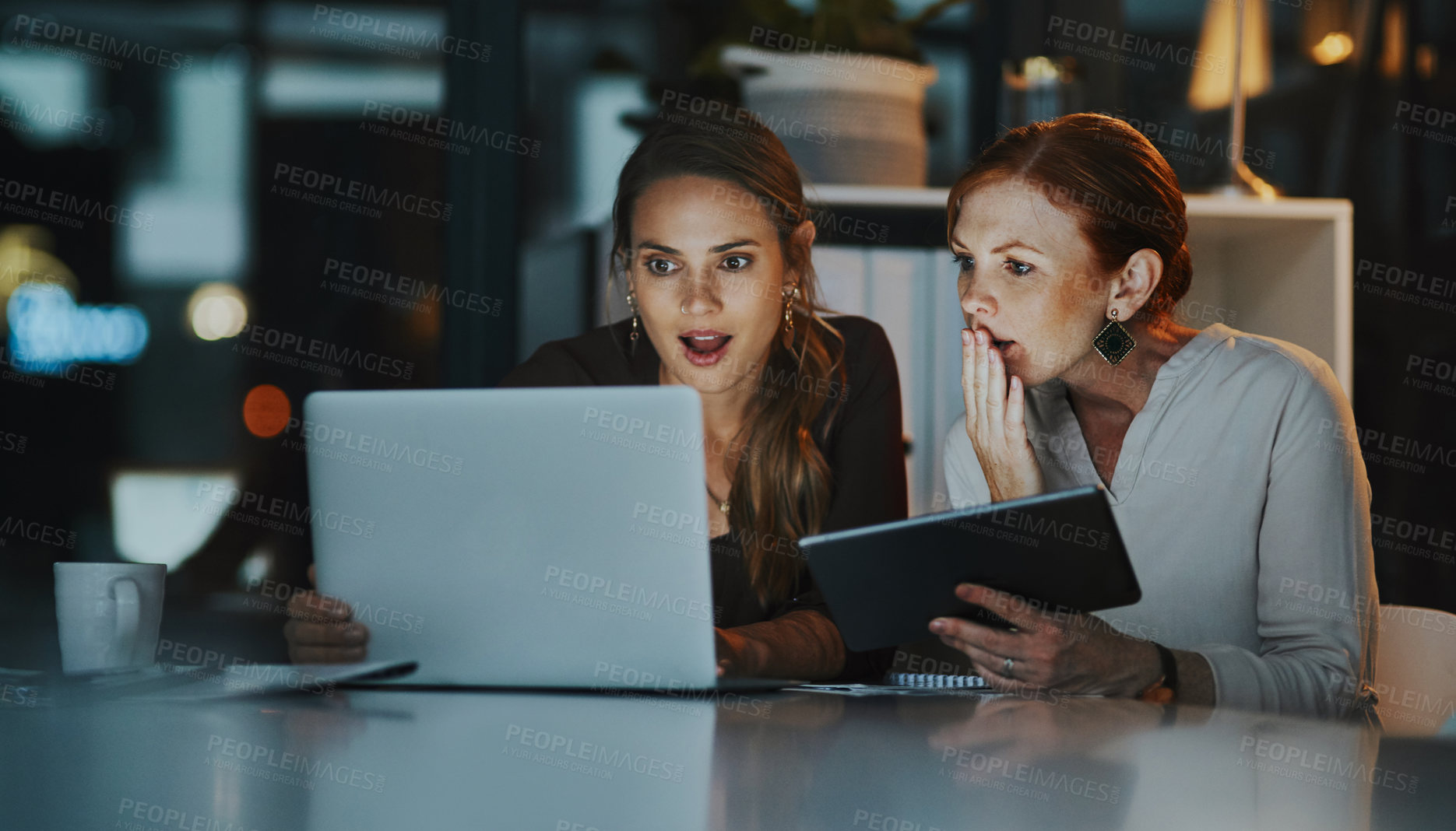 Buy stock photo Shot of two businesswomen looking surprised while working together on a laptop in an office at night
