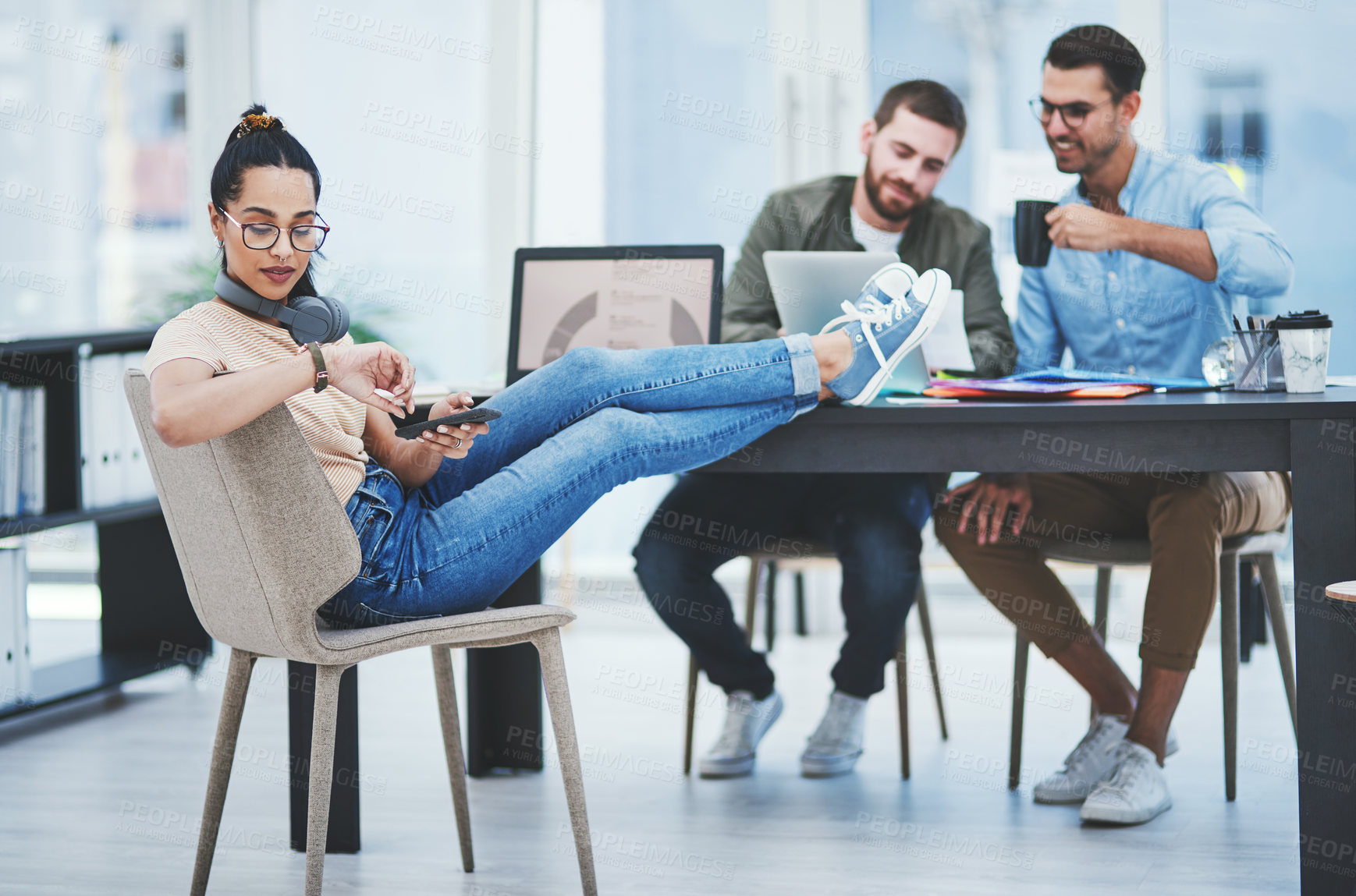 Buy stock photo Shot of a young designer checking the time while using a cellphone in an office with her colleagues in the background