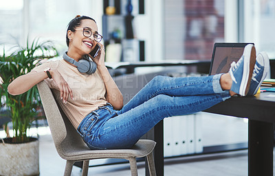 Buy stock photo Shot of a young designer talking on a cellphone while sitting with her feet up on a table in an office