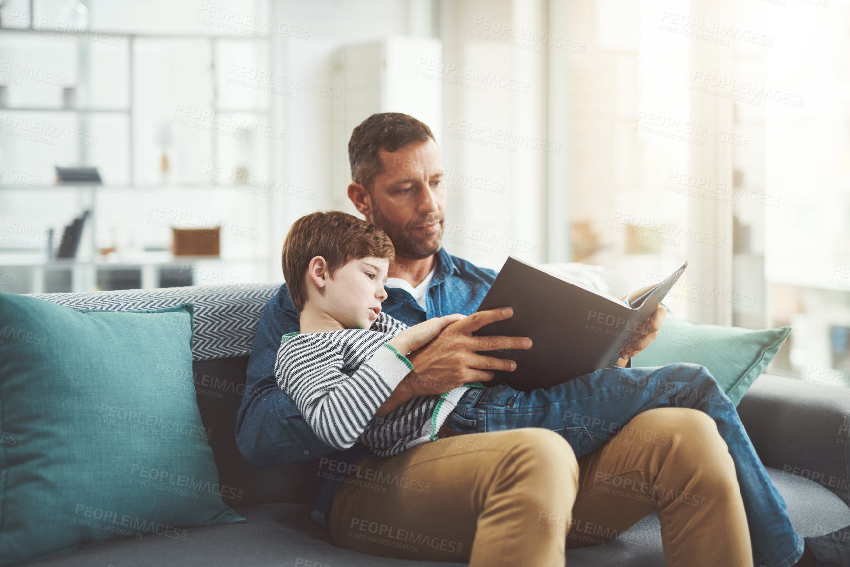 Buy stock photo Shot of a carefree little boy and his father relaxing on a sofa while reading a book at home during the day