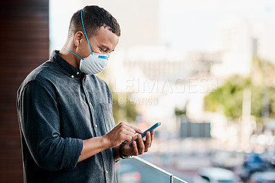 Buy stock photo Shot of a young businessman wearing a mask and using a smartphone on the balcony of an office