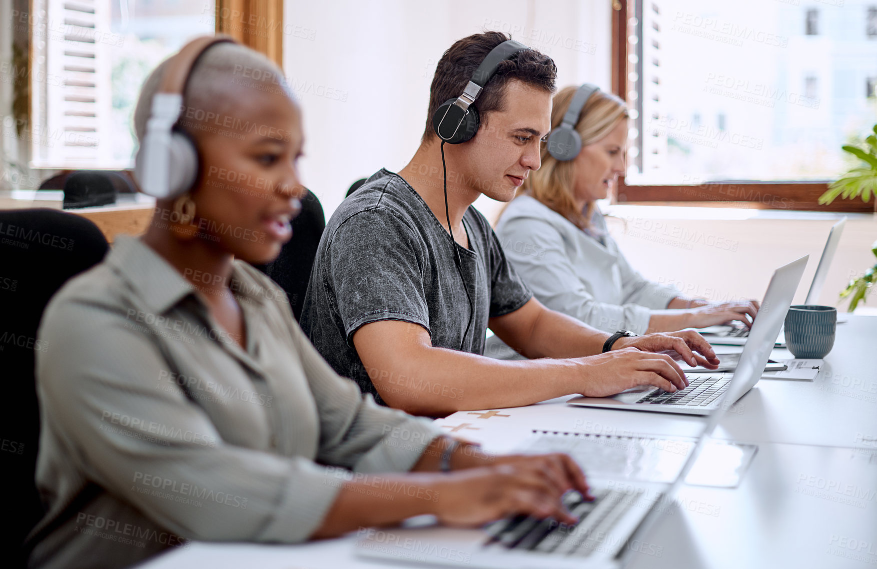 Buy stock photo Shot of a young businessman wearing headphones while working on a laptop alongside his colleagues in an office