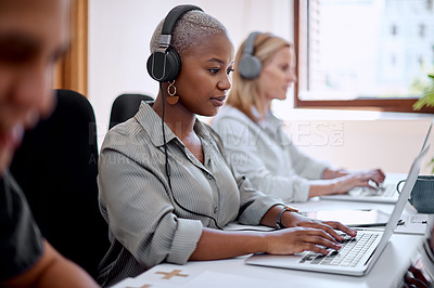 Buy stock photo Shot of a young businesswoman wearing headphones while working on a laptop alongside her colleagues in an office