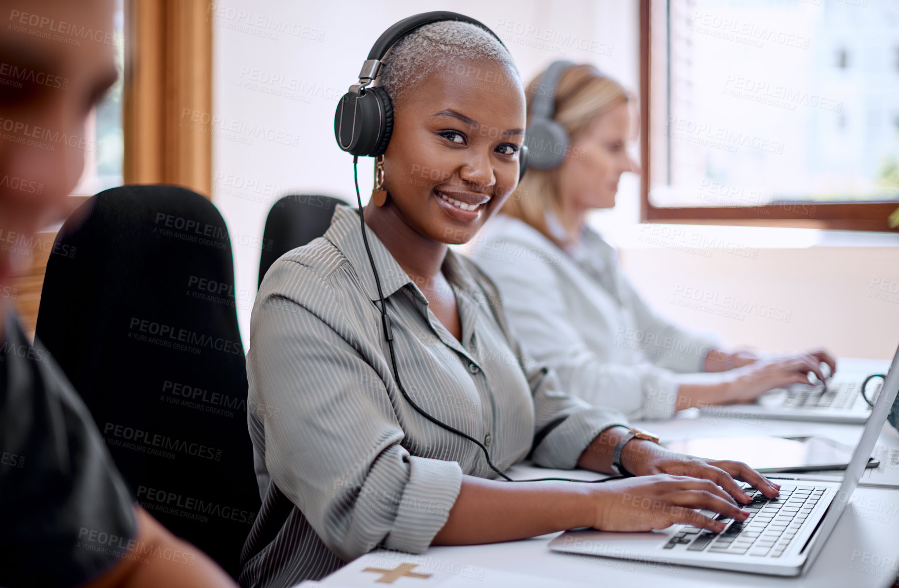 Buy stock photo Portrait of a young businesswoman wearing headphones while working on a laptop alongside her colleagues in an office