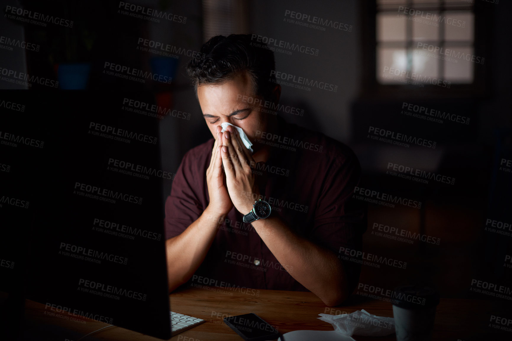 Buy stock photo Shot of a young businessman blowing his nose while working in an office at night