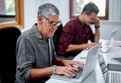 Buy stock photo Shot of a senior businessman working on a laptop in an office with a colleague in the background