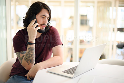 Buy stock photo Shot of a young businessman talking on a cellphone while working on a laptop in an office