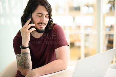 Buy stock photo Shot of a young businessman talking on a cellphone while working on a laptop in an office