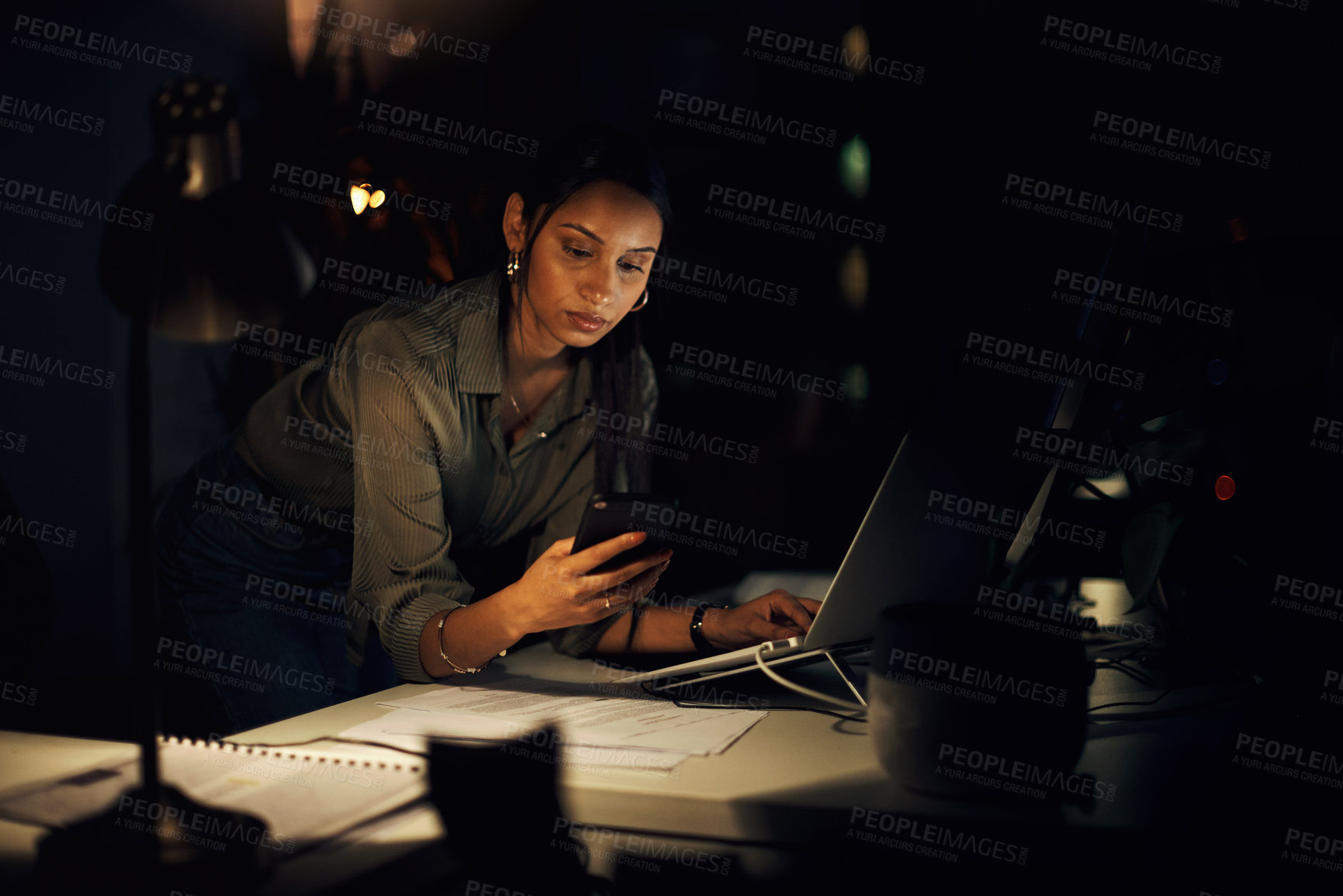 Buy stock photo Shot of a young businesswoman using a cellphone while working on a laptop in an office at night