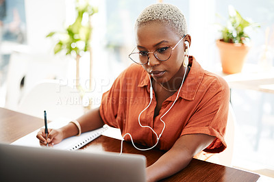 Buy stock photo Shot of a young businesswoman wearing earphones while writing notes and using a laptop in an office
