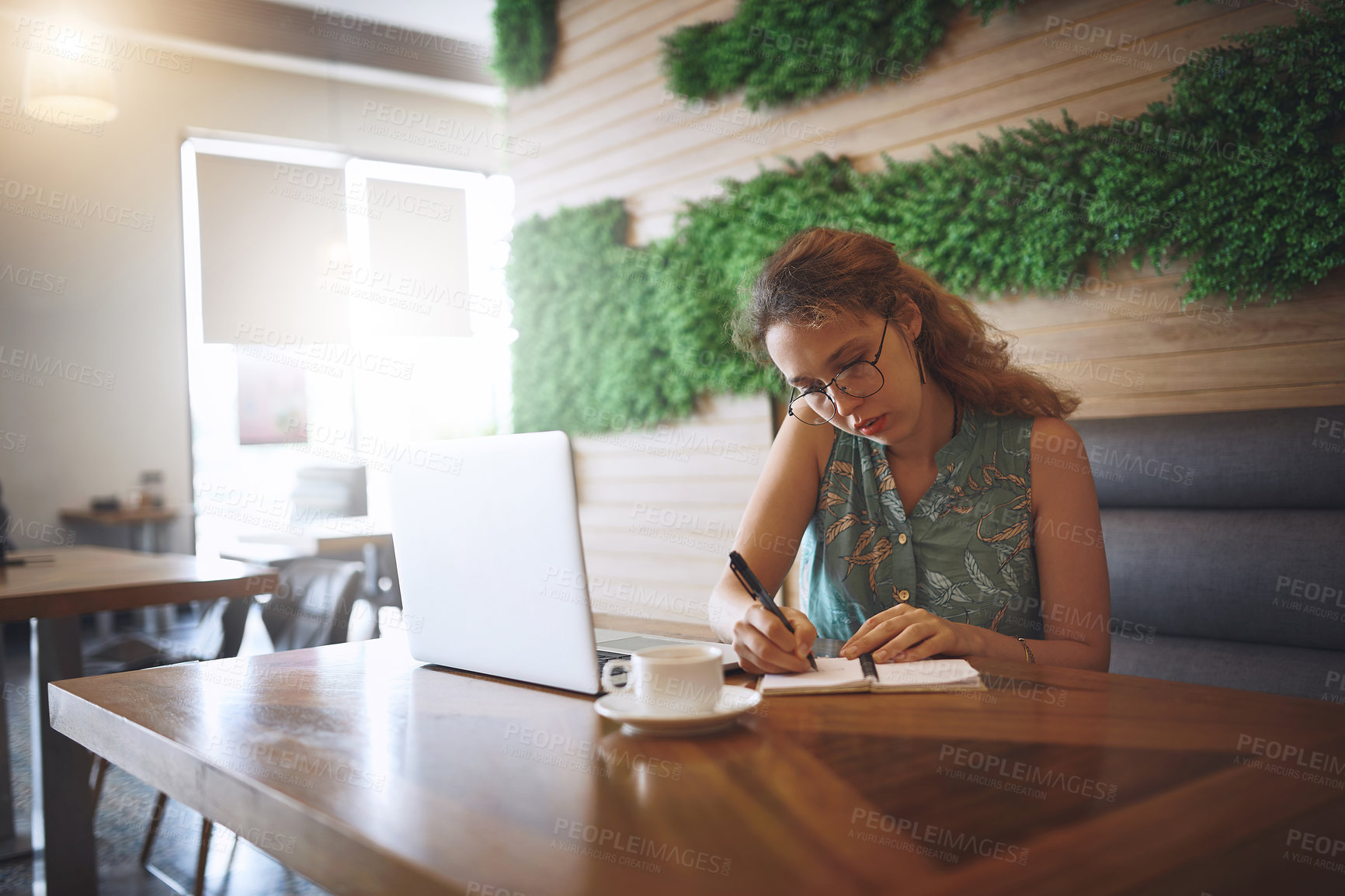 Buy stock photo Shot of a young woman using a laptop and writing in a notebook while working at a cafe