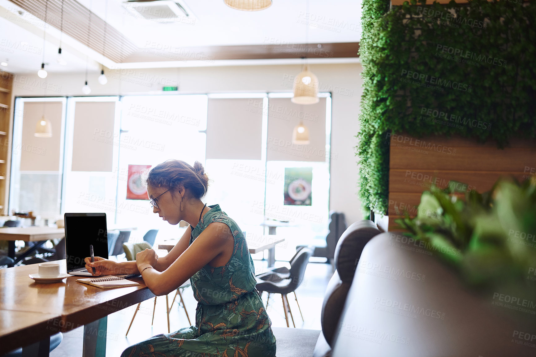 Buy stock photo Shot of a young woman using a laptop and writing in a notebook while working at a cafe