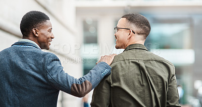 Buy stock photo Two young businessmen smiling, talking and walking through the city together, excited for their new project. Happy multiracial coworkers strolling with his hand on his friends shoulder while laughing