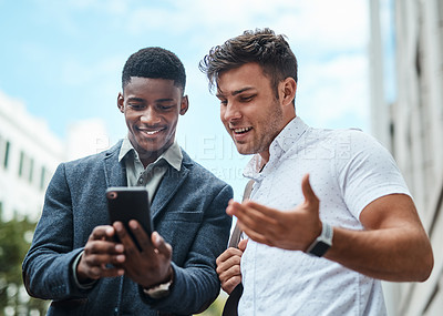 Buy stock photo Shot of two young businessmen using a smartphone together against an urban background