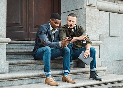 Buy stock photo Shot of two young businessmen using a smartphone together against an urban background